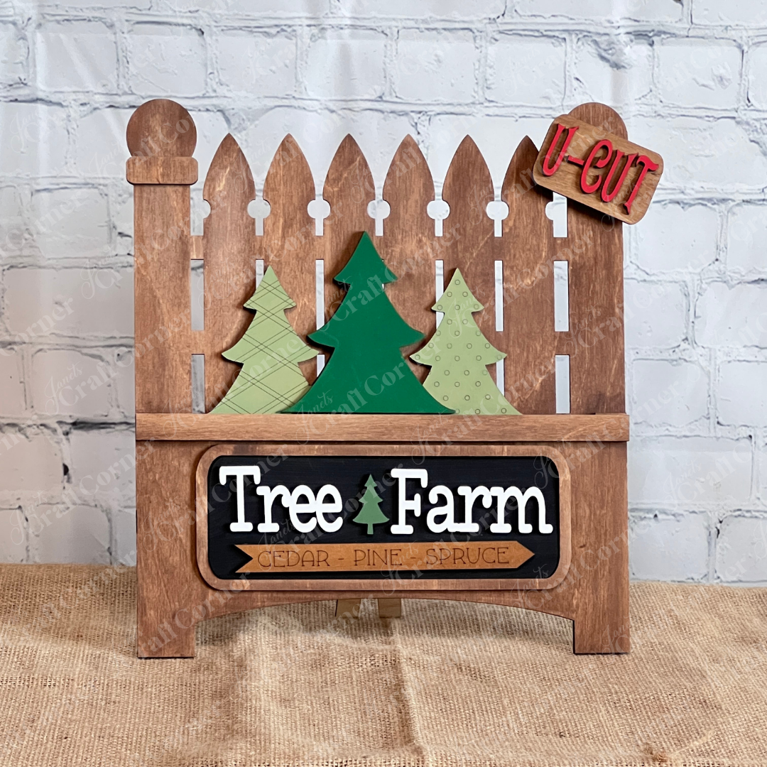 The Tree Farm Interchangeable Insert by Janet's Craft Corner is a DIY seasonal home decor kit, featuring wooden fence art with cutout Christmas trees and a "Tree Farm" sign. It includes bold red "U-Cut" letters, perfect for rustic and festive decor on a burlap surface against a white brick wall.
