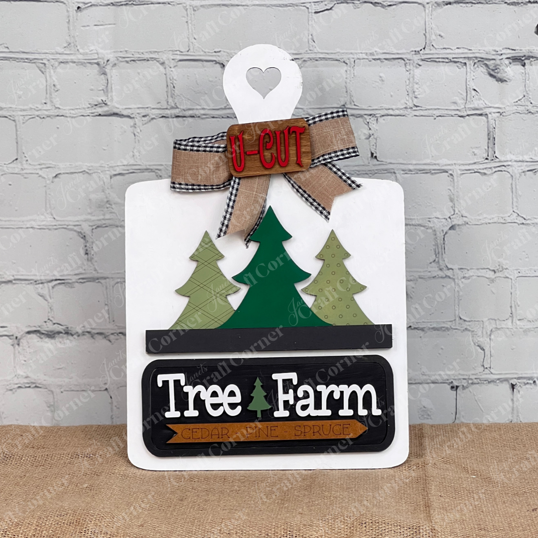 The Breadboard Interchangeable Base with Insert by Janet's Craft Corner features a decorative wooden breadboard with a heart cutout, three green trees, the text "Tree Farm" and "Cedar Pine Spruce." It's complete with a bow and "U-Cut" sign, perfect for DIY home decor against a white brick wall.