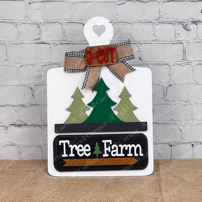 The Breadboard Interchangeable Base with Insert by Janet's Craft Corner features a decorative wooden breadboard with a heart cutout, three green trees, the text "Tree Farm" and "Cedar Pine Spruce." It's complete with a bow and "U-Cut" sign, perfect for DIY home decor against a white brick wall.