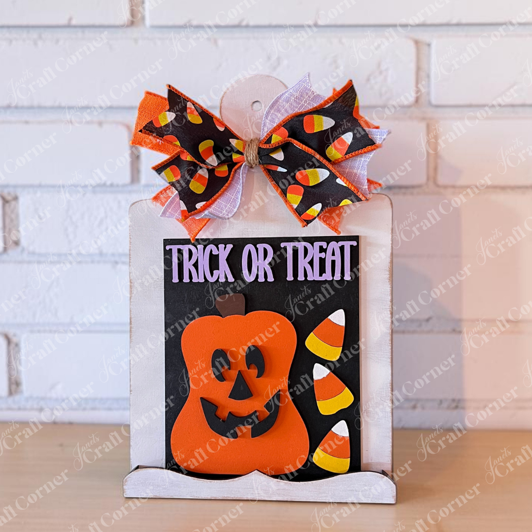 Showcase your Halloween spirit with Janet’s Craft Corner’s “Trick or Treat Pumpkin Farmhouse Sign & Breadboard Sign Holder Class,” featuring a charming sign adorned with “Trick or Treat” text, a cheerful orange pumpkin, candy corn accents, and a festive bow on top—all beautifully displayed on a rustic breadboard sign holder against a crisp white brick wall.