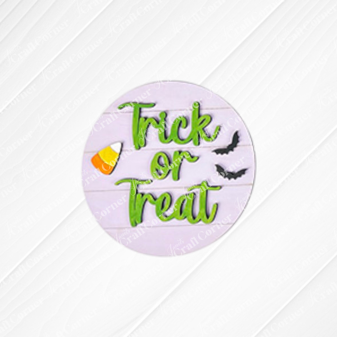 Showcase your Halloween Mini Shelf Sitters DIY kit from Janet's Craft Corner with a round sign featuring green "Trick or Treat" text, candy corn, and bats on a light wooden background—the perfect addition to your seasonal home decor.