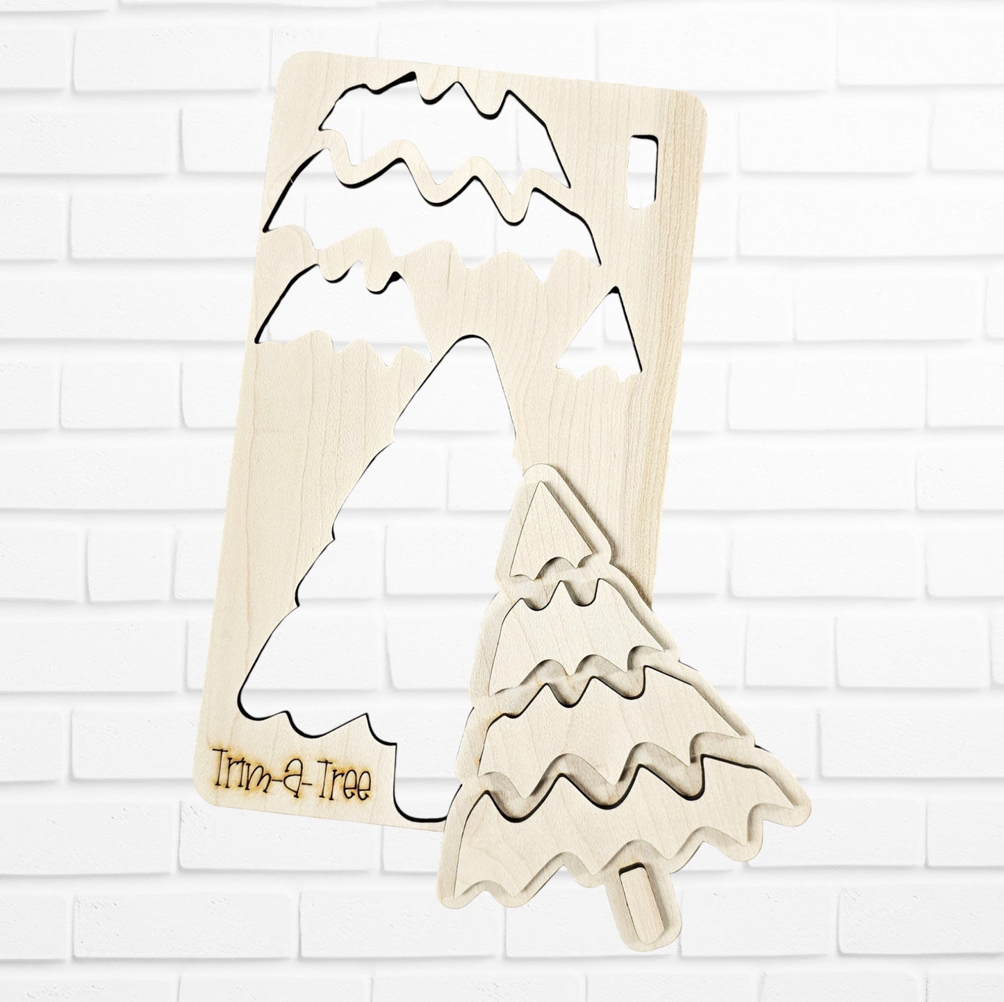 Experience the finest in holiday decor with Janet's Craft Corner's "Trim a Tree Kids Pop-Out Kit." This DIY craft kit features a wooden Christmas tree puzzle with a distinctive cut-out piece in front. The stylized tree showcases wavy branch patterns and an engraved "Trim-a-Tree" on its base. Set against a white brick wall, it serves as a charming accent for any festive setting.