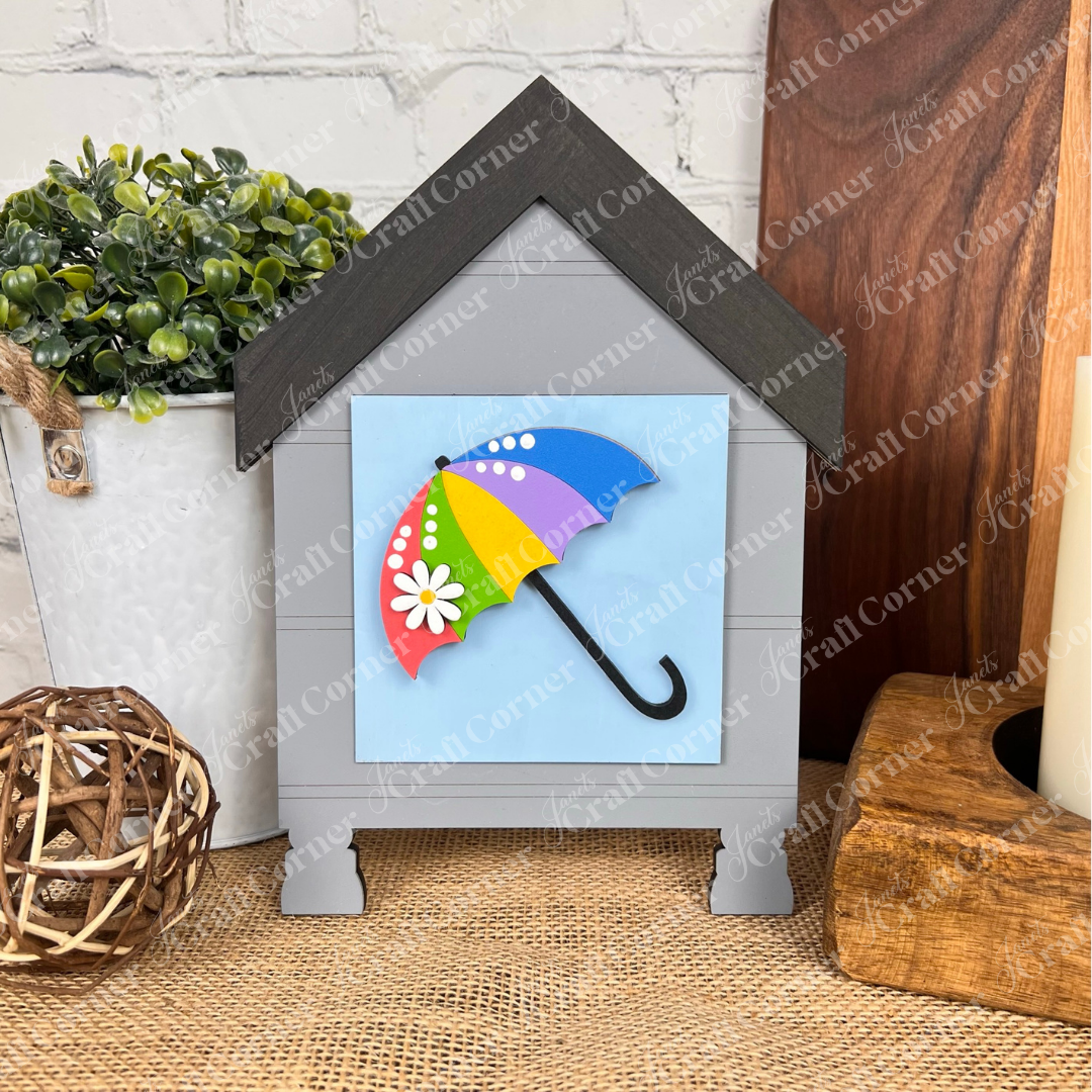 Janet's Craft Corner's "Hello Spring Squares" is a house-shaped sign featuring a daisy and colorful umbrella on blue, ideal for tiered tray decor. Displayed with a potted plant, wicker ball, and wooden items on burlap.