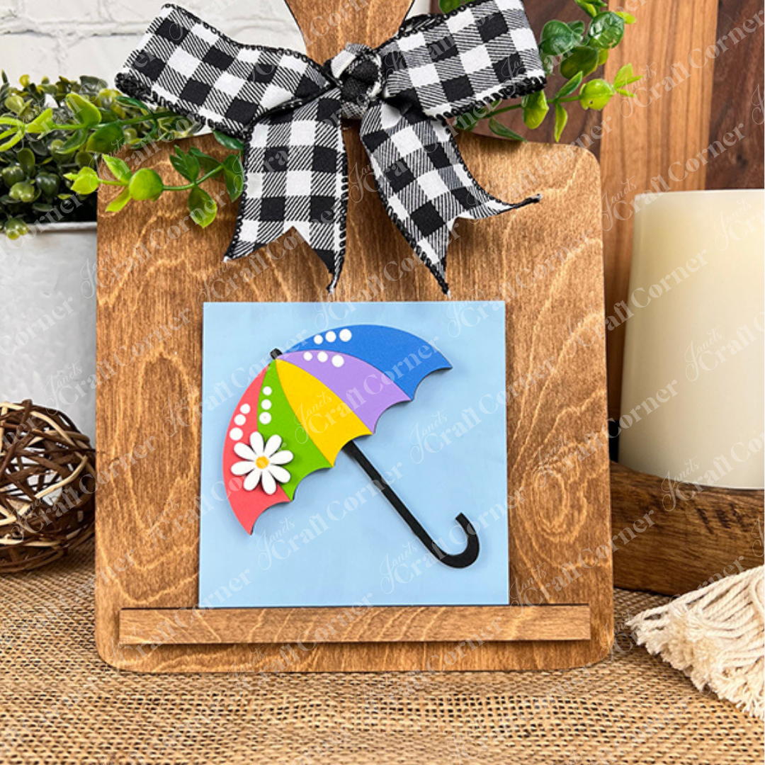 A wooden stand with a black-and-white checkered bow displays Janet's Craft Corner's Hello Spring Squares tile, featuring a colorful umbrella with daisies on a blue background. Greenery and rustic tiered tray decor add extra charm in the background.