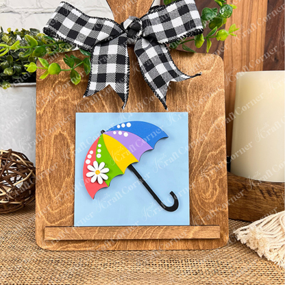 A wooden stand with a black-and-white checkered bow displays Janet's Craft Corner's Hello Spring Squares tile, featuring a colorful umbrella with daisies on a blue background. Greenery and rustic tiered tray decor add extra charm in the background.