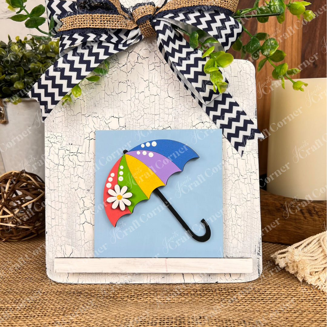 Janet's Craft Corner "Hello Spring Squares" is a small wood sign with a daisy and umbrella on a blue background, accented by a chevron bow and green foliage—perfect as tiered tray decor or with other rustic cut-outs.