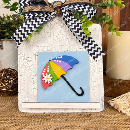 Janet's Craft Corner "Hello Spring Squares" is a small wood sign with a daisy and umbrella on a blue background, accented by a chevron bow and green foliage—perfect as tiered tray decor or with other rustic cut-outs.