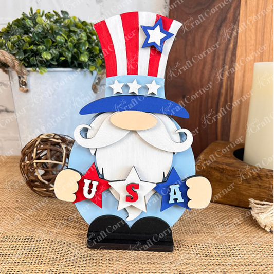 The Uncle Sam Gnome - Shelf Sitter by Janet's Craft Corner is a wooden patriotic figure featuring a charming white beard and dressed in a red and blue striped top hat accented with stars. It holds letters spelling "USA," capturing the essence of rustic farmhouse decor. Completing this delightful DIY kit theme are the background elements of a wicker ball and wooden surfaces.