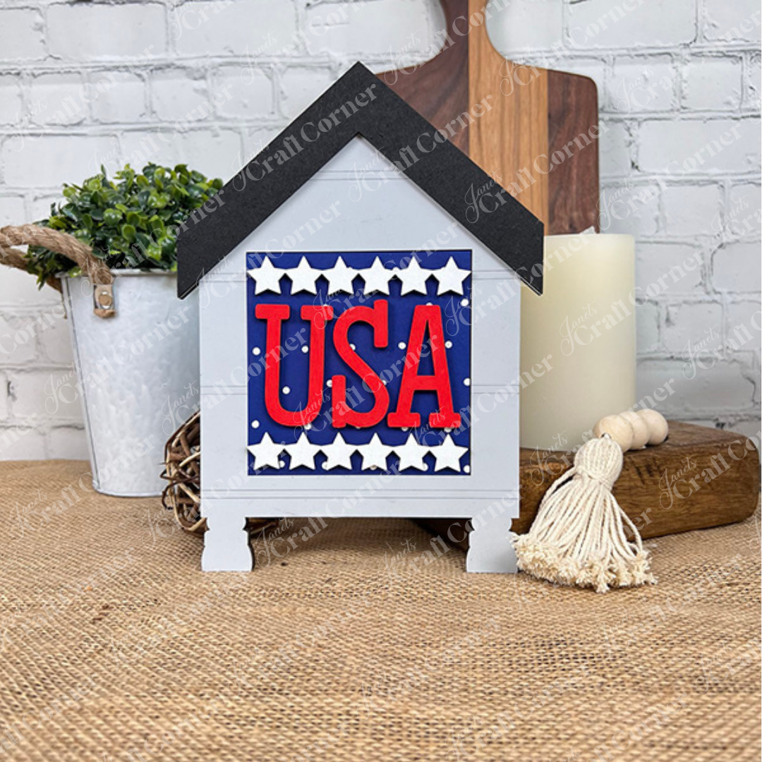 The Patriotic Squares DIY craft kit by Janet's Craft Corner is a house-shaped sign with bold red "USA" letters on blue and white stars—perfect for farmhouse-style Independence Day decor on tabletop or against white brick walls.