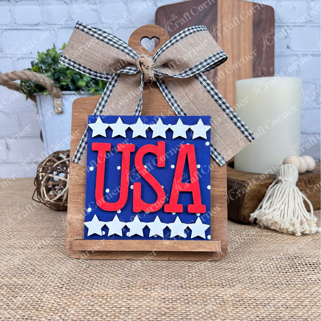 The Patriotic Squares DIY craft kit from Janet's Craft Corner features a small wooden sign with "USA" in bold red on a blue, starry background. Ideal for Independence Day décor, it complements rustic accents like candles and greenery.