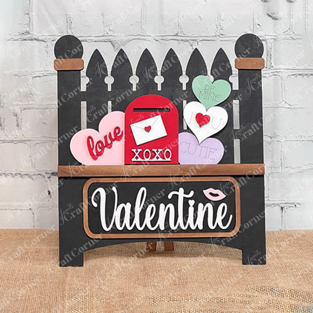 The Valentine Interchangeable Insert by Janet's Craft Corner is a DIY home decor kit featuring a black picket fence with heart-shaped messages, a red "XOXO" mailbox, "Valentine" text, and an interchangeable insert for customization.
