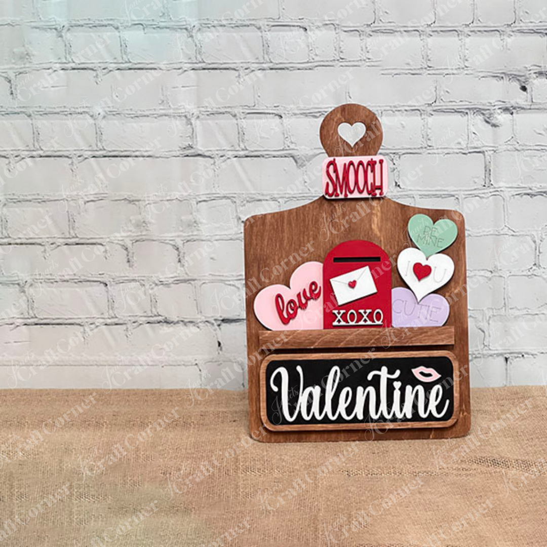 Janet's Craft Corner offers a Valentine Interchangeable Insert DIY home decor kit featuring a wooden board with a heart cutout, colorful hearts with messages like "love" and "cutie," an envelope, and a blackboard with "Valentine," perfect for creating personalized Valentine's Day displays.