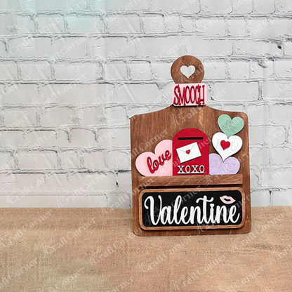 Janet's Craft Corner offers a Valentine Interchangeable Insert DIY home decor kit featuring a wooden board with a heart cutout, colorful hearts with messages like "love" and "cutie," an envelope, and a blackboard with "Valentine," perfect for creating personalized Valentine's Day displays.