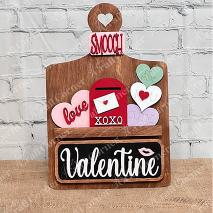 Janet's Craft Corner offers a DIY home decor kit called the Breadboard Interchangeable Base with Insert. It includes a wooden base shaped like a cutting board, featuring "Smooch" on top, interchangeable accessories, and designs like hearts with words like "Be Mine," plus "XOXO" and "Valentine.