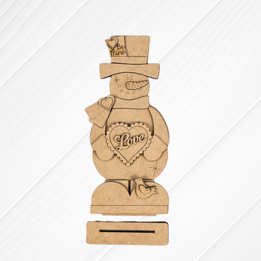 The Valentine Snowman Shelf Sitter by Janet's Craft Corner is a DIY decor craft kit that includes a wooden snowman with heart-shaped "Love" and "Be Mine" details, on a rectangular base with a small bird figure against a textured white and burlap background. Perfect for Valentine's Day decorating.