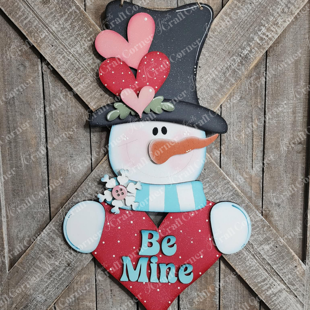The Valentine Snowman Door Hanger kit by Janet’s Craft Corner features a carrot-nosed snowman with red and pink hearts, a blue scarf, and black hat holding a “Be Mine” heart—perfect DIY seasonal home decor against rustic wooden planks. Set of 1.