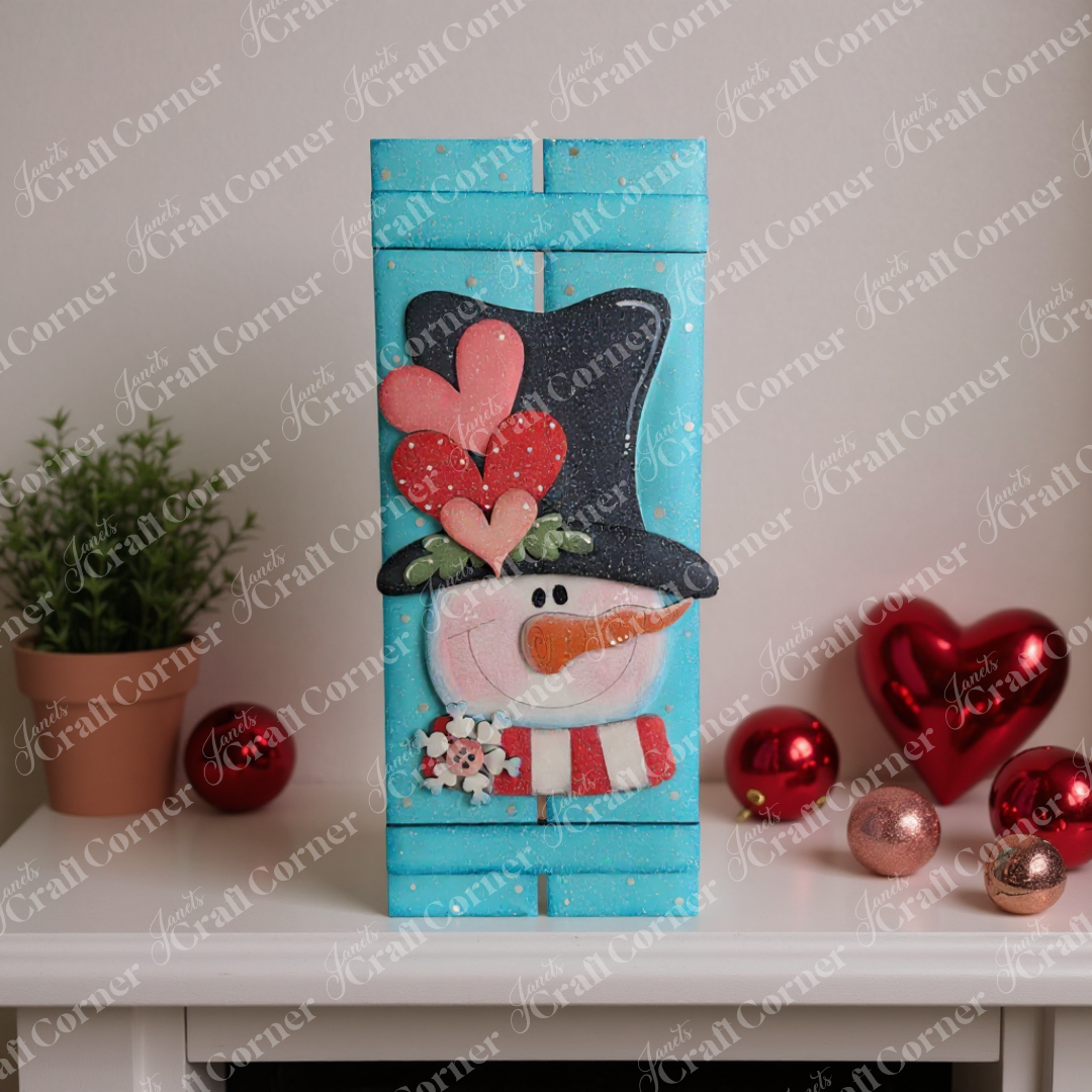 The Valentine Snowman Table Top Pallet Shelf Leaner by Janet's Craft Corner is a DIY seasonal home decor kit featuring a snowman with heart accents, perfect for adding winter charm to your shelf alongside plants and ornaments.