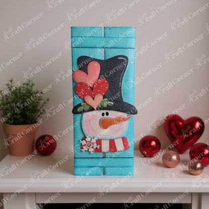 The Valentine Snowman Table Top Pallet Shelf Leaner by Janet's Craft Corner is a DIY seasonal home decor kit featuring a snowman with heart accents, perfect for adding winter charm to your shelf alongside plants and ornaments.