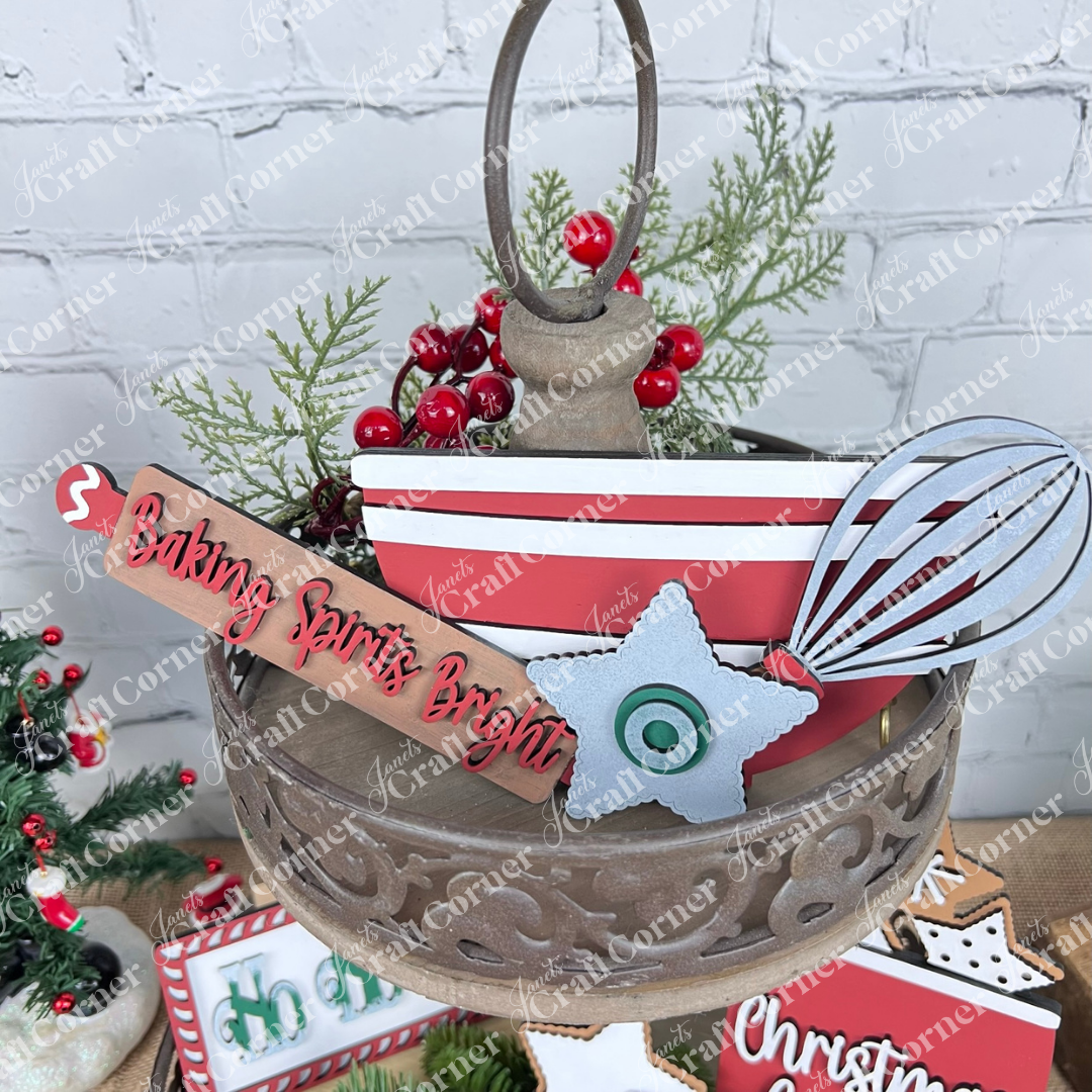 A rustic metal tiered tray displays festive red and white decor, featuring Janet's Craft Corner Vintage Santa Cookies Decor Set DIY kit, a "Baking Spirits Bright" sign, whisk, faux greenery, berries, and a star cookie ornament.