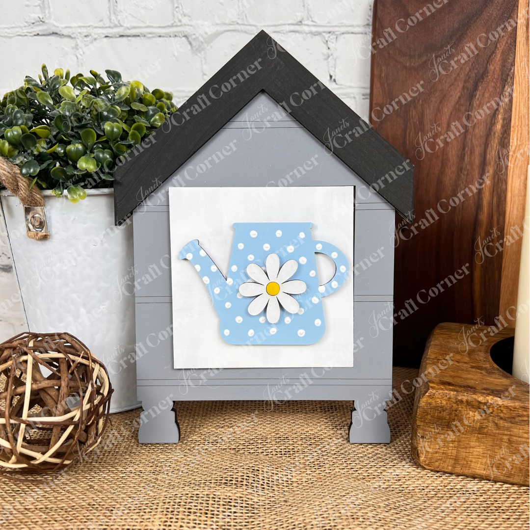 Janet's Craft Corner Bloom & Grow Squares: A gray house-shaped wood sign with black roof, blue polka-dot watering can, and white daisy. Ideal for tiered tray decor beside a potted plant and wicker ball.