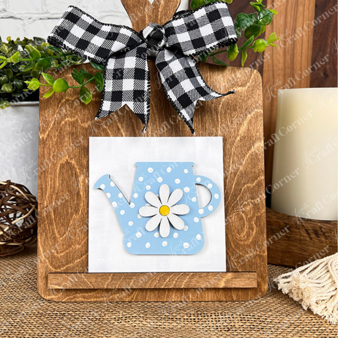 Janet’s Craft Corner’s Bloom & Grow Squares features a white tile with a blue polka-dot watering can and daisy design on a wooden cutting board with a checkered bow—ideal tiered tray decor with greenery accents.