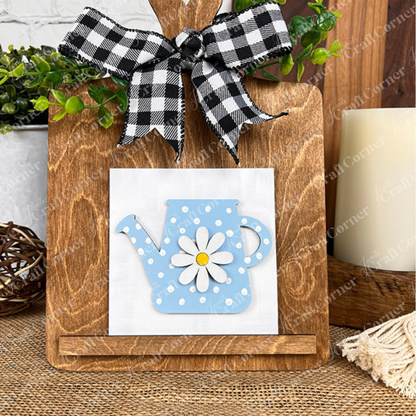 Janet’s Craft Corner’s Bloom & Grow Squares features a white tile with a blue polka-dot watering can and daisy design on a wooden cutting board with a checkered bow—ideal tiered tray decor with greenery accents.