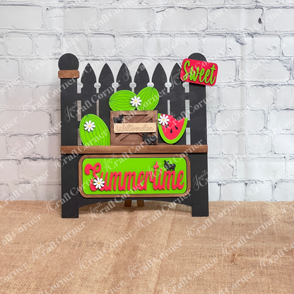 Introducing the **Summertime Watermelon Interchangeable Insert - Painted** by *Janet's Craft Corner*: This hand-painted decoration on a black garden fence base showcases watermelons and a watermelon slice, paired with a green "Summertime" sign. Enhanced with black silhouettes of ants and white daisy flowers, this charming piece is perfect for your home decor.