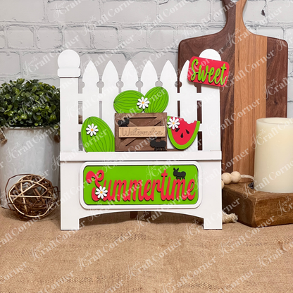 Introducing the **Summertime Watermelon Interchangeable Insert - Painted** by *Janet's Craft Corner*: This hand-painted decoration on a white garden fence base showcases watermelons and a watermelon slice, paired with a green "Summertime" sign. Enhanced with black silhouettes of ants and white daisy flowers, this charming piece is perfect for your home decor.