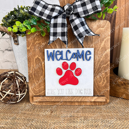 Janet's Craft Corner Dog Squares features a wooden stand with a black-and-white plaid bow, blue "Welcome," red paw print, and "Hope you like dog hair," accented by greenery and tiered tray decor in the background.