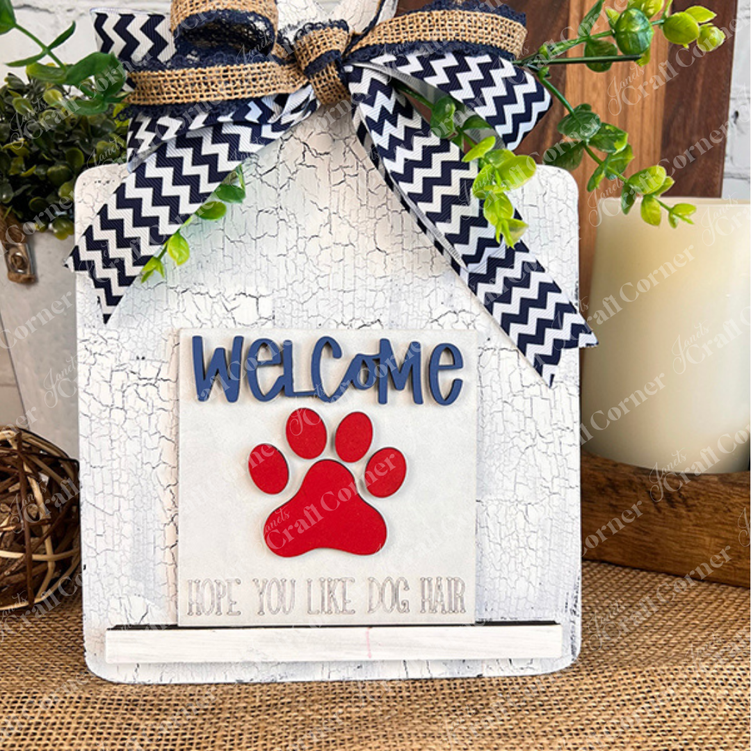 Janet's Craft Corner Dog Squares is a decorative sign for tiered trays, featuring a blue and white chevron ribbon, green foliage, a large red paw print, and "WELCOME Hope you like dog hair" on rustic cracked white paint.