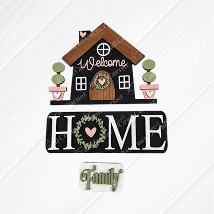 Introducing the "Welcome Home Interchangeable Insert," a DIY home decor kit by Janet's Craft Corner. This decorative insert features a house design with hand-painted lettering, including "Welcome" at the top and "HOME" adorned with a heart-shaped wreath in the "O," followed by the word "Family." The background showcases a charming burlap and white quilted appearance, making it ideal for versatile home decor.