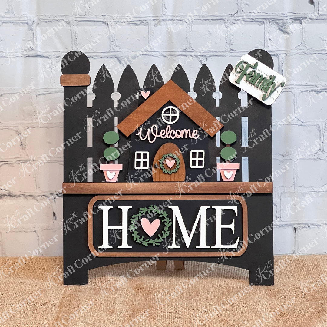 The Welcome Home Interchangeable Insert by Janet's Craft Corner is a DIY home decor kit featuring a charming  black wooden picket fence sign with a central house, "Welcome" on top, "HOME" below. It includes inserts with potted plants and signs reading "Family" beside a heart.