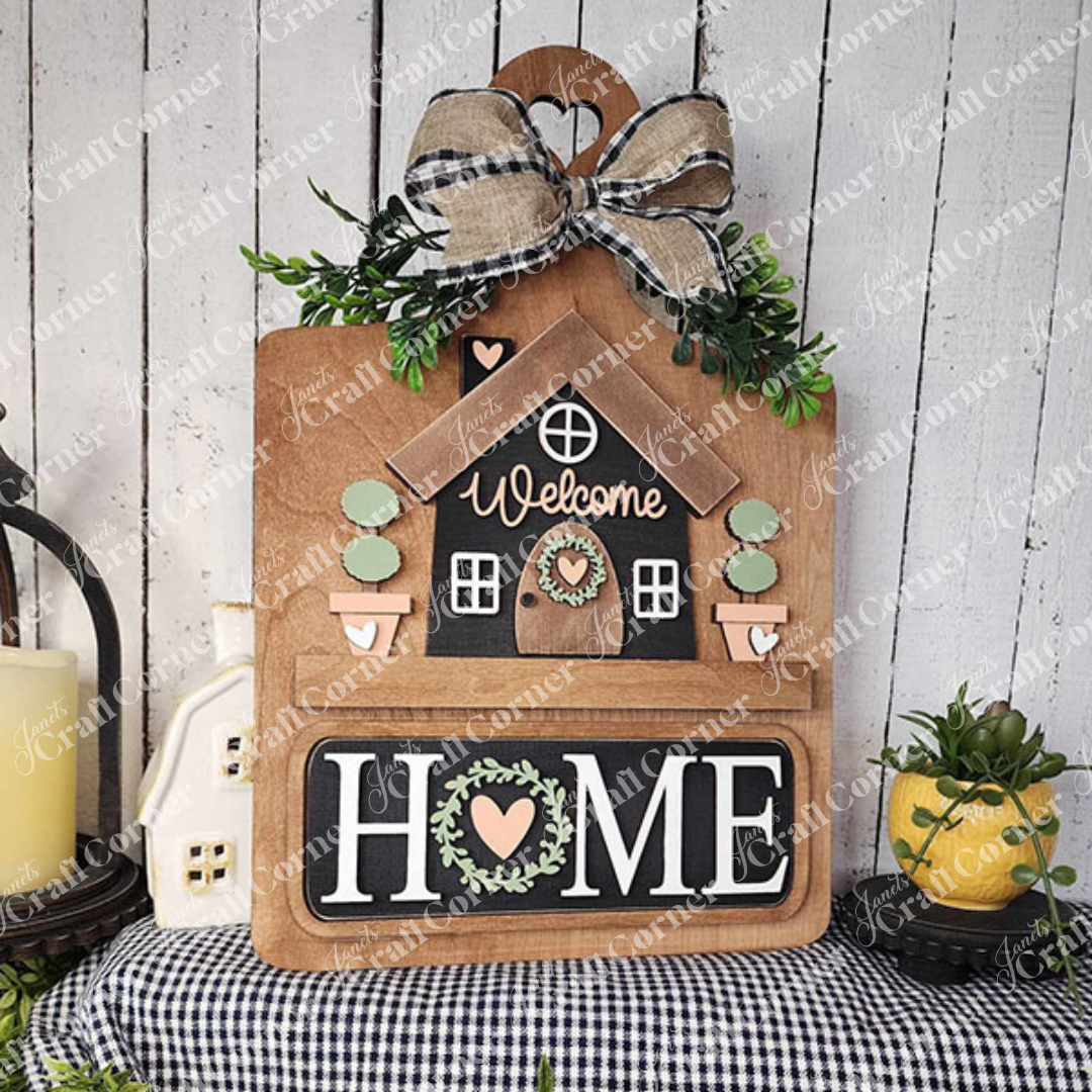 The Welcome Home Interchangeable Insert from Janet's Craft Corner is a DIY home decor kit featuring a house design with a "Welcome" message, adorned with an elegant plaid bow and greenery. The bold "HOME" letters are highlighted with a heart and wreath symbol, set against a rustic backdrop for versatile home decoration.