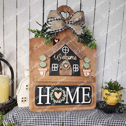 The Welcome Home Interchangeable Insert from Janet's Craft Corner is a DIY home decor kit featuring a house design with a "Welcome" message, adorned with an elegant plaid bow and greenery. The bold "HOME" letters are highlighted with a heart and wreath symbol, set against a rustic backdrop for versatile home decoration.