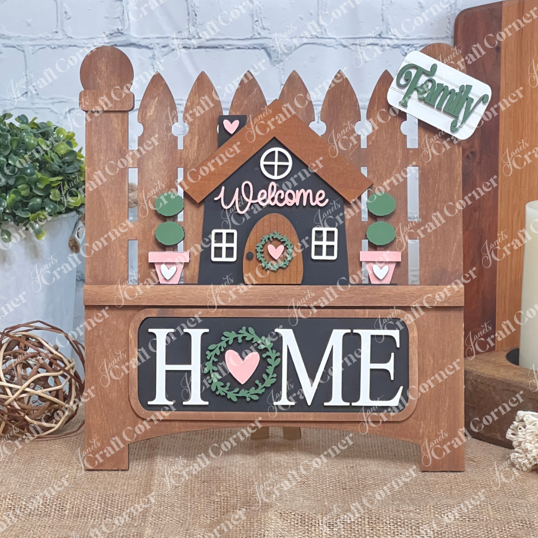 The Welcome Home Interchangeable Insert by Janet's Craft Corner is a DIY home decor kit featuring a charming stained wooden picket fence sign with a central house, "Welcome" on top, "HOME" below. It includes inserts with potted plants and signs reading "Family" beside a heart.