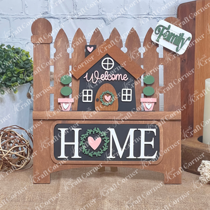 The Welcome Home Interchangeable Insert by Janet's Craft Corner is a DIY home decor kit featuring a charming stained wooden picket fence sign with a central house, "Welcome" on top, "HOME" below. It includes inserts with potted plants and signs reading "Family" beside a heart.