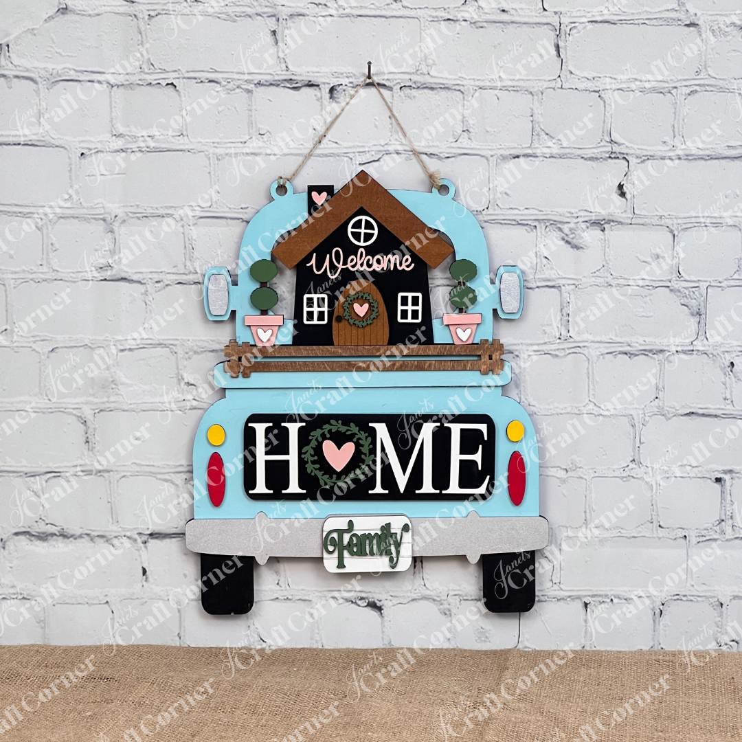 Janet's Craft Corner presents the "Welcome Home Interchangeable Insert" DIY kit. This set includes a blue truck sign with a house and "Welcome" sign, featuring "HOME" on the tailgate with a heart-shaped "O," and a mini family sign, ideal for an inviting farmhouse decor against white brick walls.