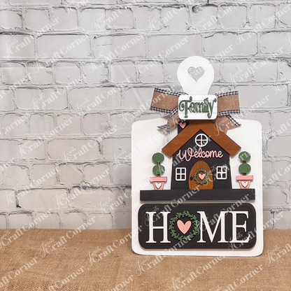 The Welcome Home Interchangeable Insert from Janet's Craft Corner is a DIY home decor kit featuring a house design with a "Welcome" message, adorned with an elegant plaid bow and greenery. The bold "HOME" letters are highlighted with a heart and wreath symbol, displayed on a white breadboard base and set against a rustic backdrop for versatile home decoration.