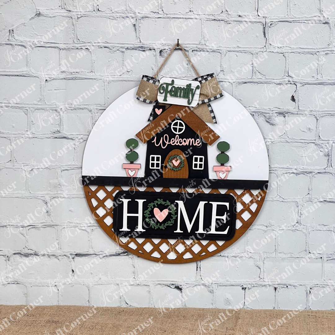 Janet's Craft Corner offers the "Welcome Home Interchangeable Insert," a DIY home decor craft kit featuring a charming round wall piece with a house, "Welcome," and phrases like "Family" and "Home" on a white brick background with heart motifs and greenery. Perfect for enhancing any space.