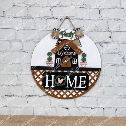 Janet's Craft Corner offers the "Welcome Home Interchangeable Insert," a DIY home decor craft kit featuring a charming round wall piece with a house, "Welcome," and phrases like "Family" and "Home" on a white brick background with heart motifs and greenery. Perfect for enhancing any space.