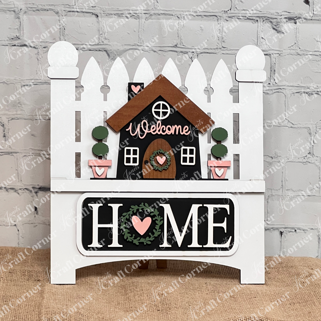 This charming farmhouse decor from Janet's Craft Corner features a small wooden house with "Welcome" atop, set against a white picket fence. Below, "HOME" with a heart inside the "O" rests on burlap against a light brick wall. Offered as the Welcome Home Interchangeable Insert DIY craft kit.