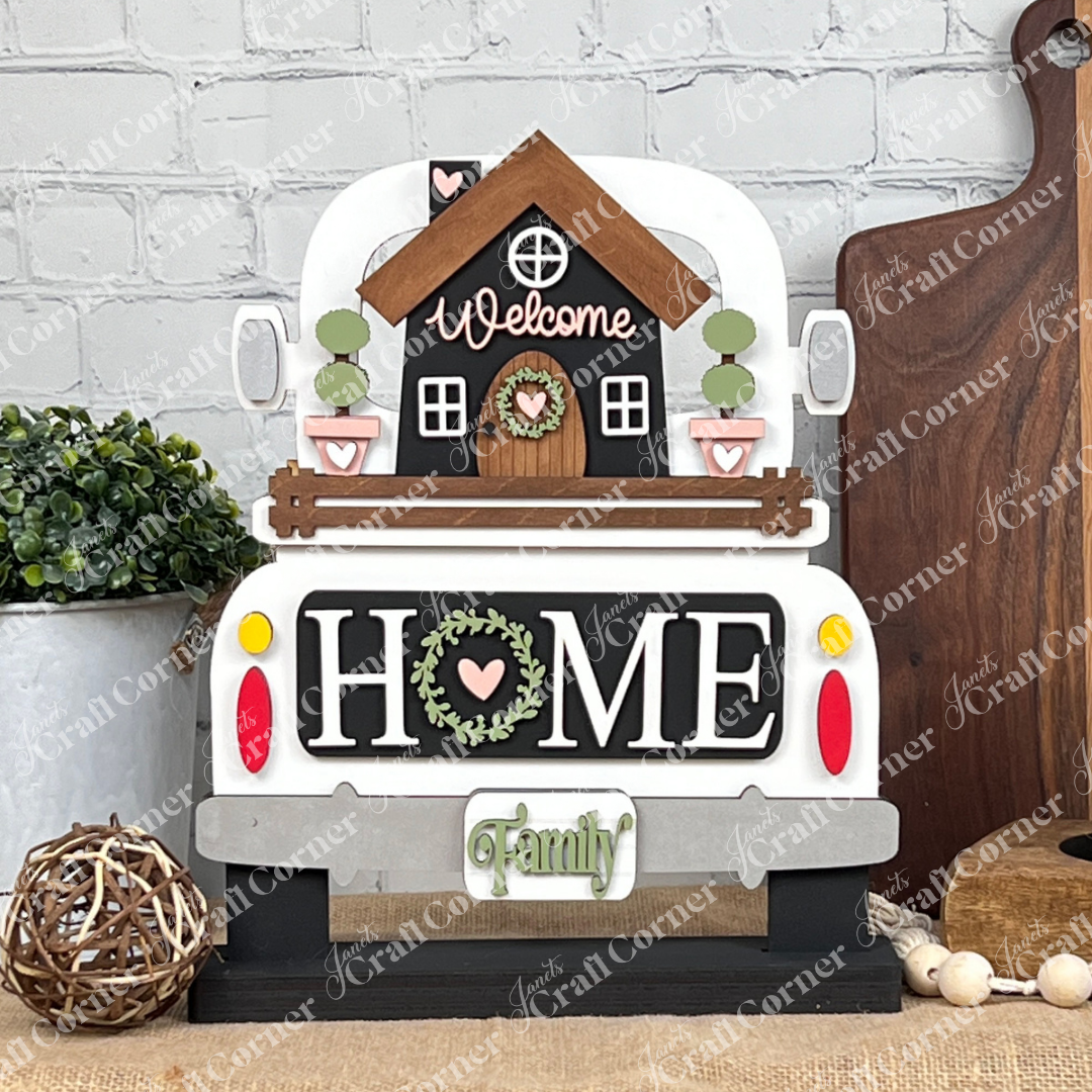 A decorative setup from Janet's Craft Corner showcases the DIY home decor kit, "Welcome Home Interchangeable Insert," featuring a hand-painted wooden truck sign highlighting the words "Welcome" and "Home." The display is enhanced with a potted plant, a wooden cutting board, a candle on a stand, and a woven ball against the backdrop of a brick wall. This customizable touch adds unique charm to any space.