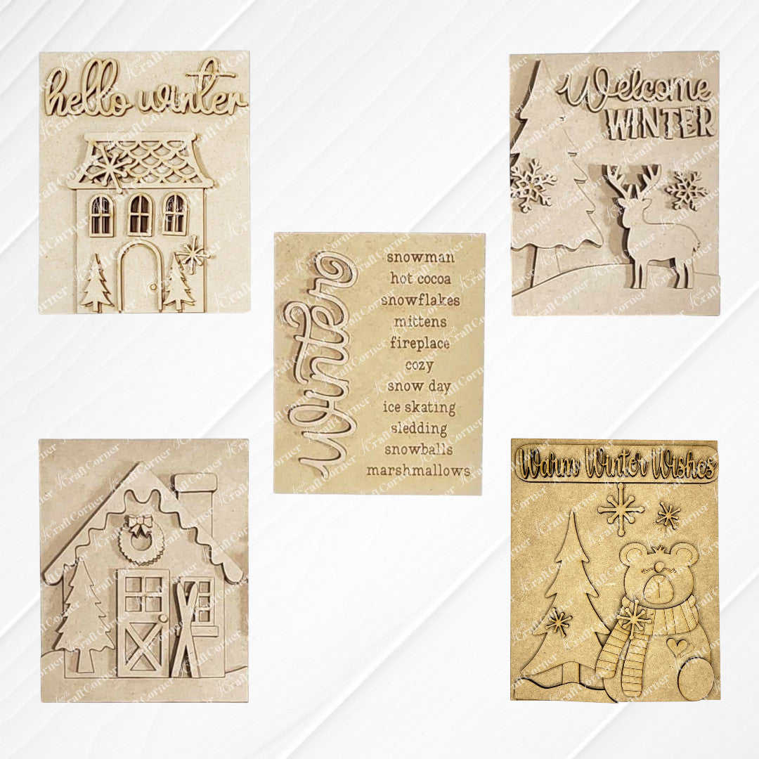 Displayed on a white wood backdrop are three winter-themed decorations: Janet's Craft Corner's Welcome Winter farmhouse signs featuring a deer, a polar bear, a list of winter words, a Victorian house and a house adorned with snow and skis. These items make for perfect home decor pieces.