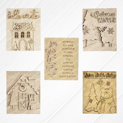 Displayed on a white wood backdrop are three winter-themed decorations: Janet's Craft Corner's Welcome Winter farmhouse signs featuring a deer, a polar bear, a list of winter words, a Victorian house and a house adorned with snow and skis. These items make for perfect home decor pieces.
