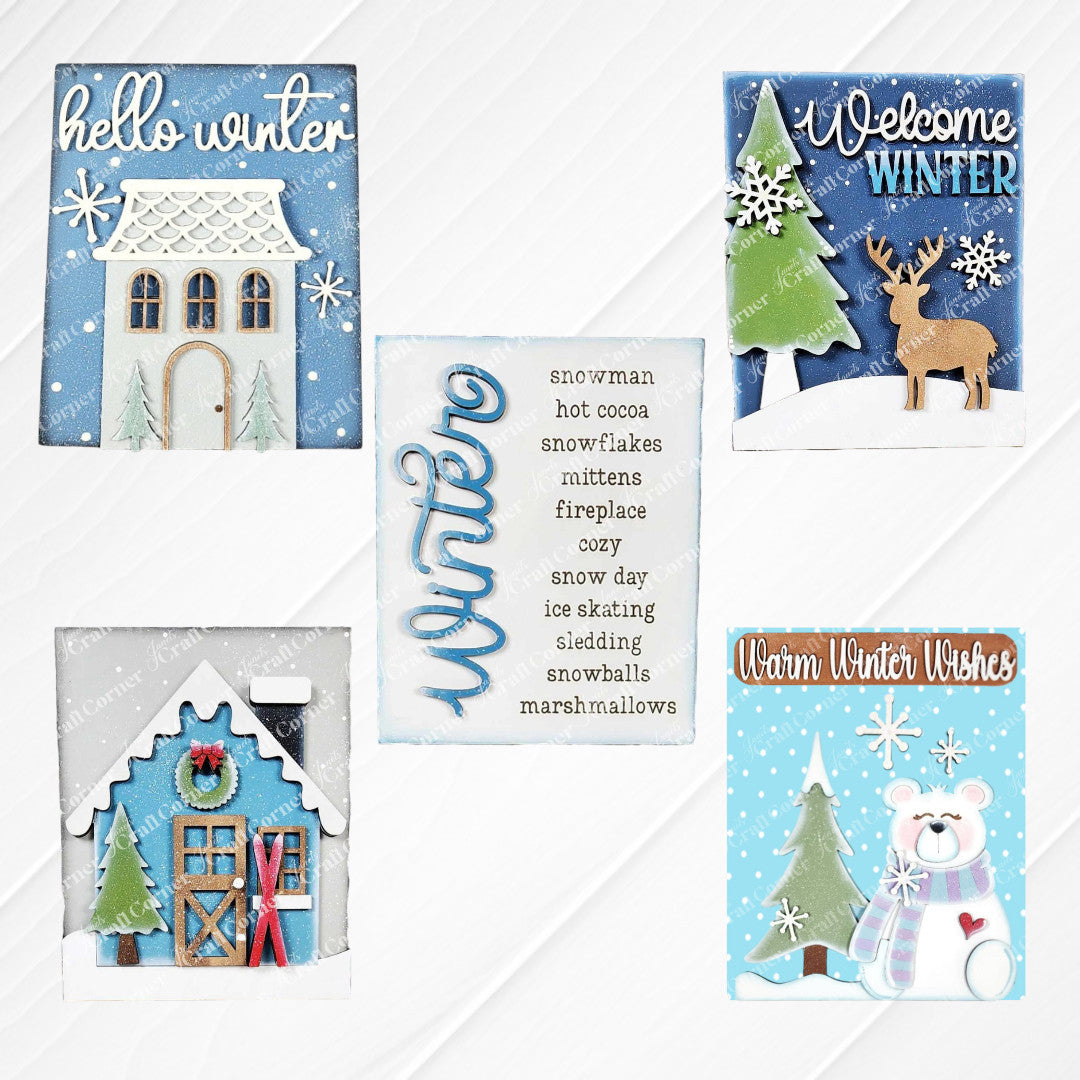 Displayed on a white wood backdrop are five winter-themed decorations: Janet's Craft Corner's Welcome Winter farmhouse signs featuring a deer, a polar bear, a list of winter words, a Victorian house and a house adorned with snow and skis. Items are shown painted and make for perfect home decor pieces.