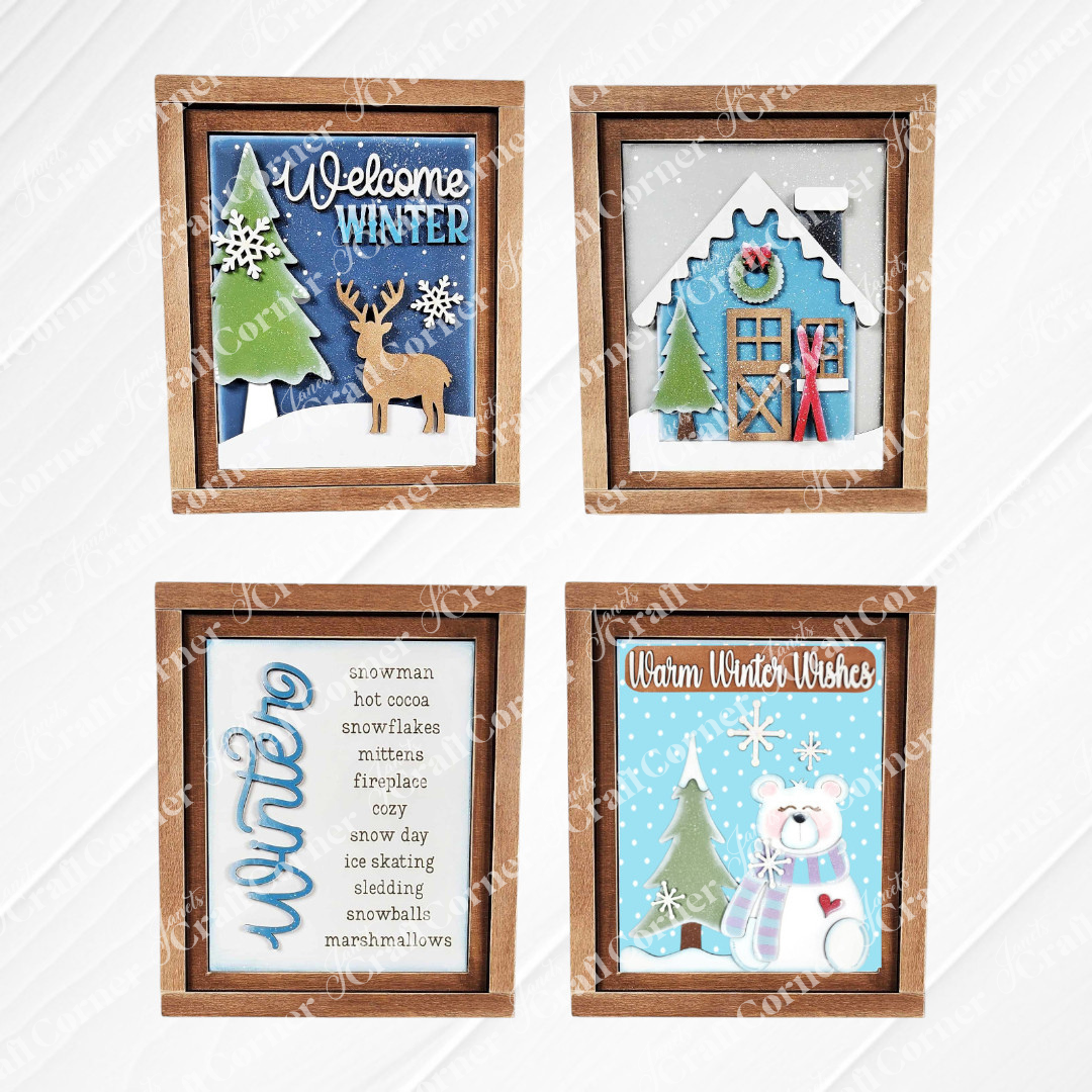 Displayed against a white wooden backdrop are four winter-themed decorations: Janet's Craft Corner's hand-painted Welcome Winter farmhouse signs featuring a deer, a polar bear, a framed list of winter words, and a house adorned with snow and skis. These items make for perfect home decor pieces.