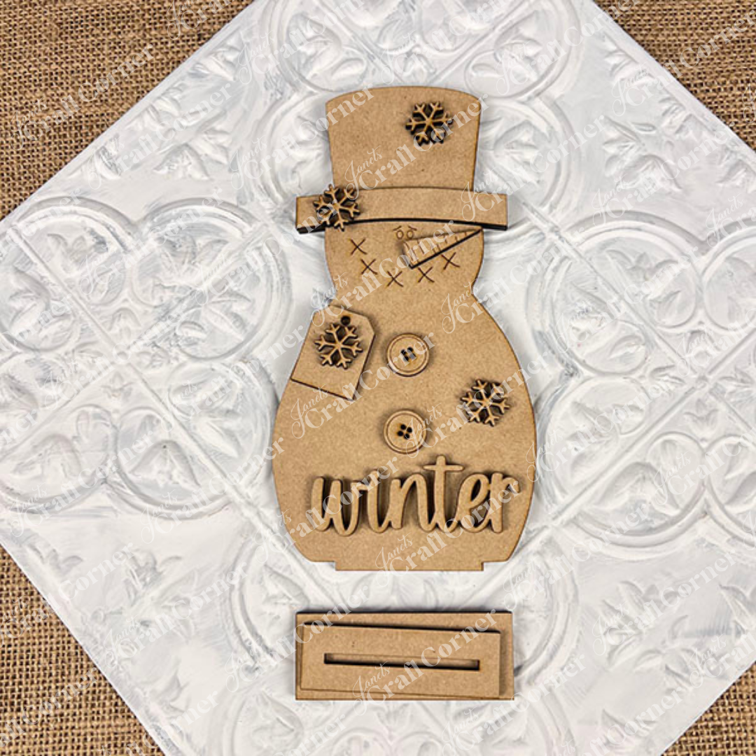 The Janet’s Craft Corner Snowman Shelf Sitters feature button and snowflake accents, “winter” at the base, and a stand—ideal festive décor for your home, displayed on a textured white square backdrop.