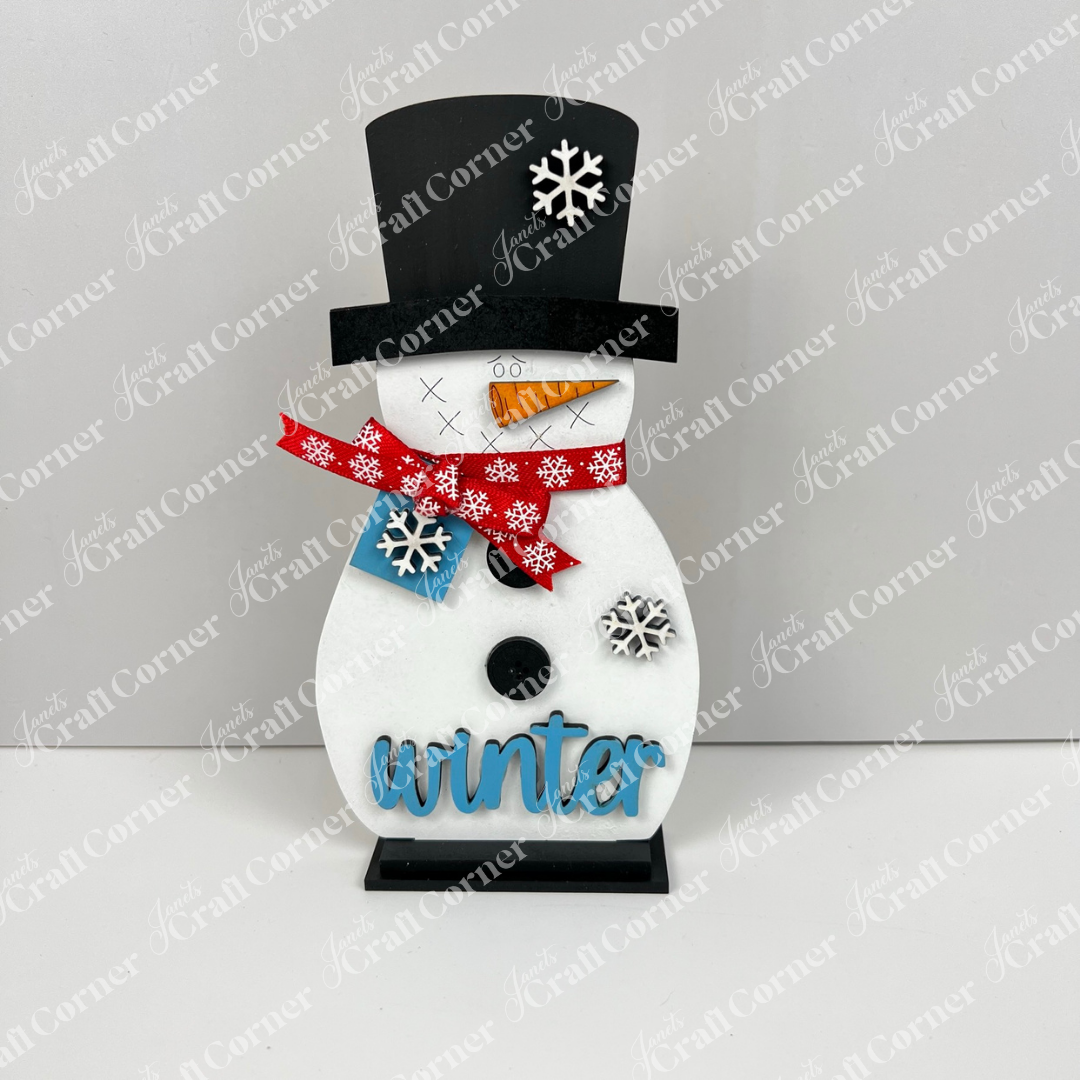 Janet's Craft Corner Snowman Shelf Sitters feature a black top hat with white snowflakes, red scarf, carrot nose, blue buttons, and "winter" in blue. The sturdy black base makes it perfect for festive winter home decor.