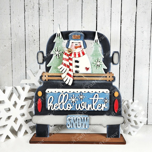 Janet's Craft Corner Hello Winter Snowman Interchangeable Insert is a DIY seasonal home decor kit featuring a blue truck, snowman, trees, and "hello winter"/"SNOW" signs on a white wood background with wooden snowflakes. Kit includes 3 pieces.