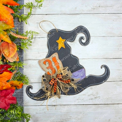 Janet’s Craft Corner Witch Hat Wall Hanger—a black wall decoration featuring a star, the number 31, bows, and autumn leaves—rests on a white wooden surface beside orange and green foliage, making it an ideal DIY wood craft kit for creating personalized Halloween decor.
